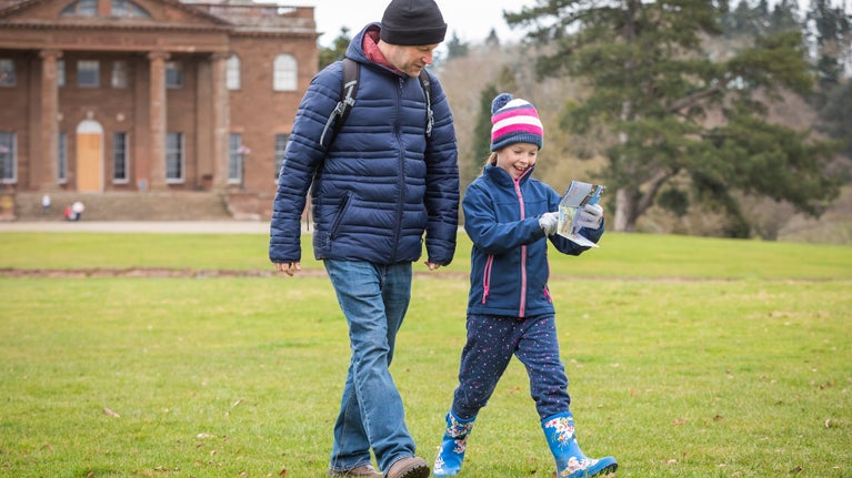 A man and a girl walking while looking at a map, dressed in winter clothes and wellies, with Berrington Hall in the background